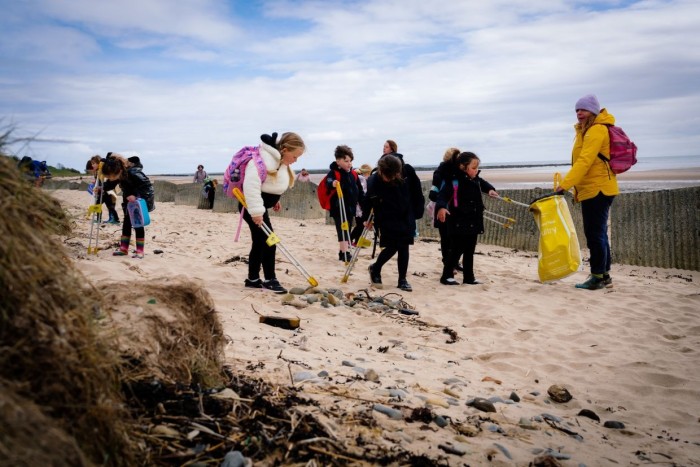 Children and adults taking part in a beach clean-up, using litter pickers and bags to collect rubbish from a sandy beach on a cloudy day.
