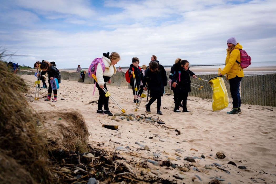 Children and adults taking part in a beach clean-up, using litter pickers and bags to collect rubbish from a sandy beach on a cloudy day.